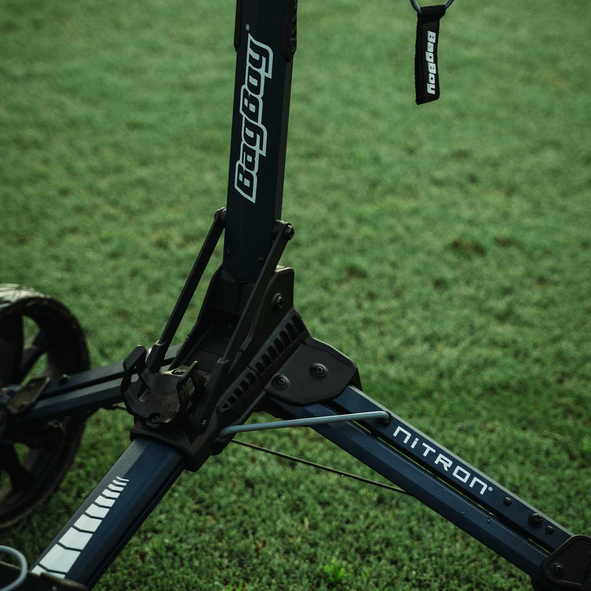 Detailed close-up of the Bag Boy branding on the frame of the Nitron golf push cart, highlighting the premium charcoal and navy colorway and rugged construction on a lush green golf course.